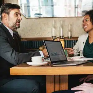 Gerente imobiliário sentado a mesa com um notebook juntamente com outros 2 corretores em um clima de conversa amistosa