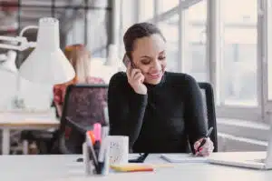 Mulher negra, de cabelo preso e um lindo sorriso no rosto trabalhando como assistente imobiliário. Ao telefone em uma ligação e fazendo anotações. Ao lado tem uma caneca e na frente um notebook branco.